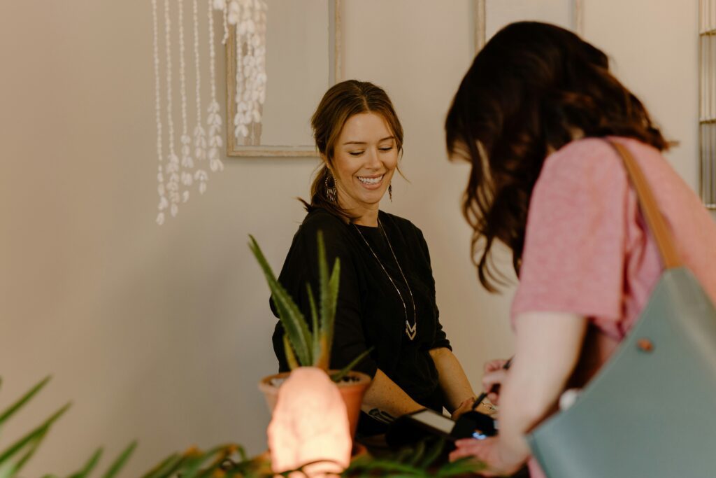 A smiling female receptionist or shop assistant with tied-back brown hair and a black top watches a customer from behind as they use a card machine on the counter. An aloe vera plant and a lit pink salt lamp are on the counter.