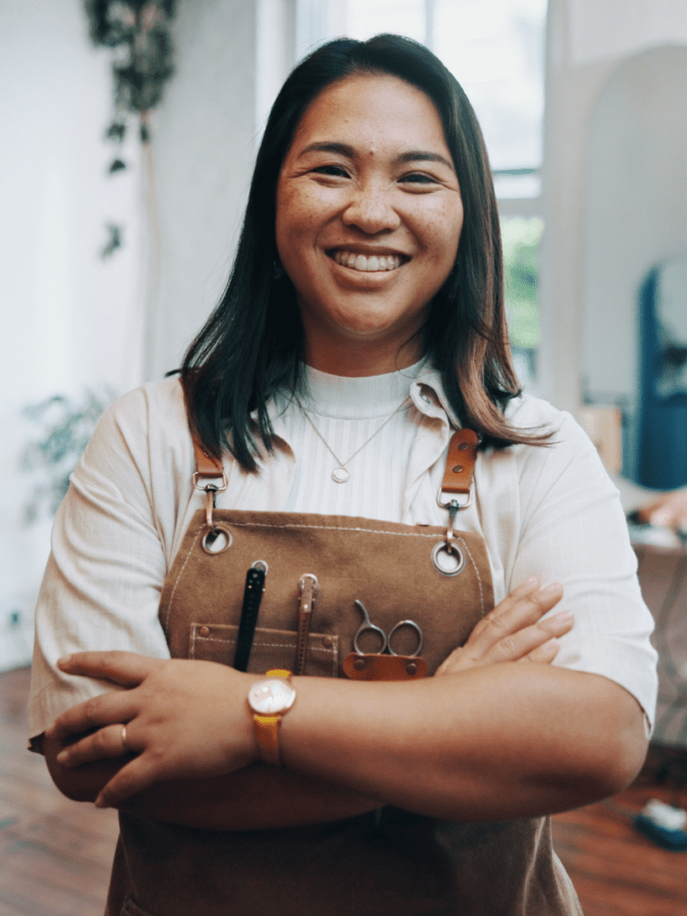 A portrait of a smiling female salon professional with dark hair, looking directly at the camera with her arms crossed. She is wearing a light-coloured shirt under a brown canvas apron equipped with hairdressing clips and scissors.
