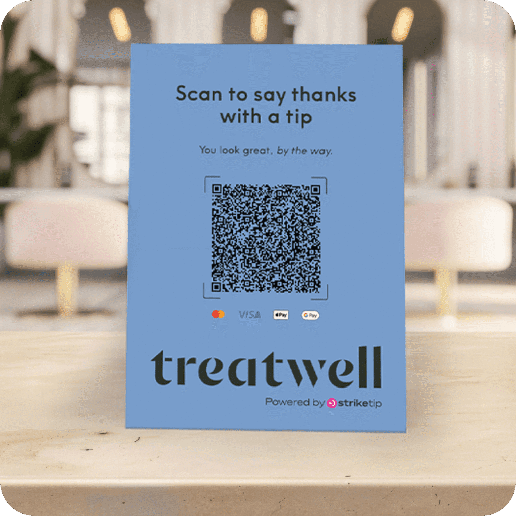 A light blue desk sign standing on a wooden counter in a blurred salon setting. The sign features a large QR code and the text "Scan to say thanks with a tip", with the Treatwell and StrikeTip logos at the bottom.