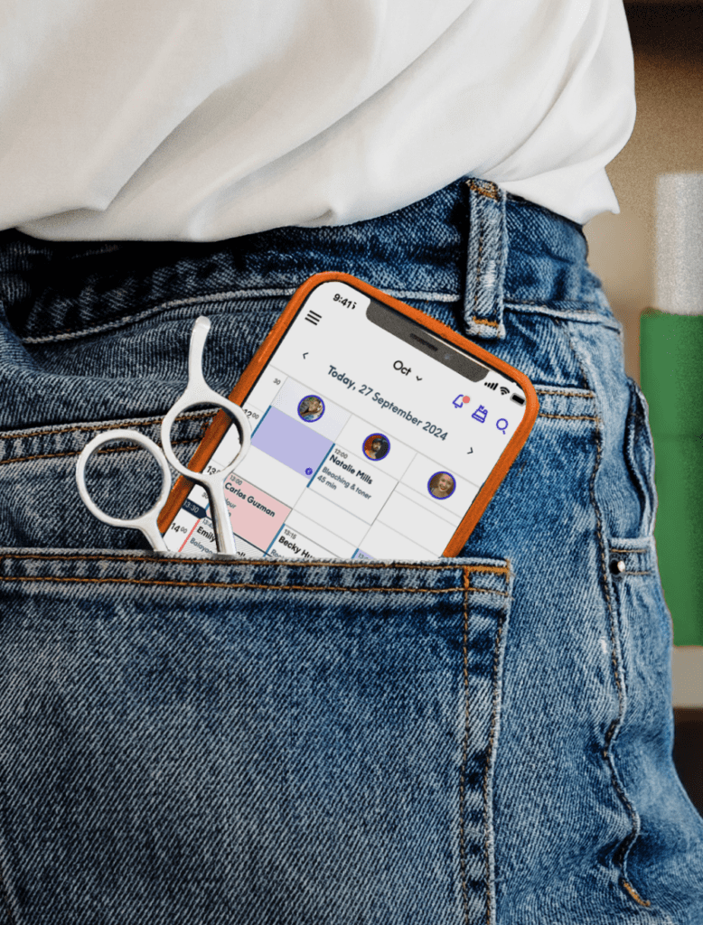 A smartphone and hairdressing scissors tucked into a jeans back pocket, the screen showing the Treatwell app's booking calendar with a "new appointment" notification.