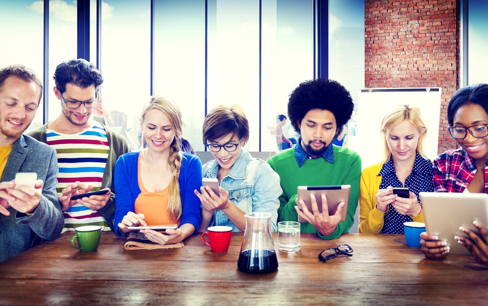 A diverse group of hair and beauty professionals sitting together at a table, using smartphones and tablets.