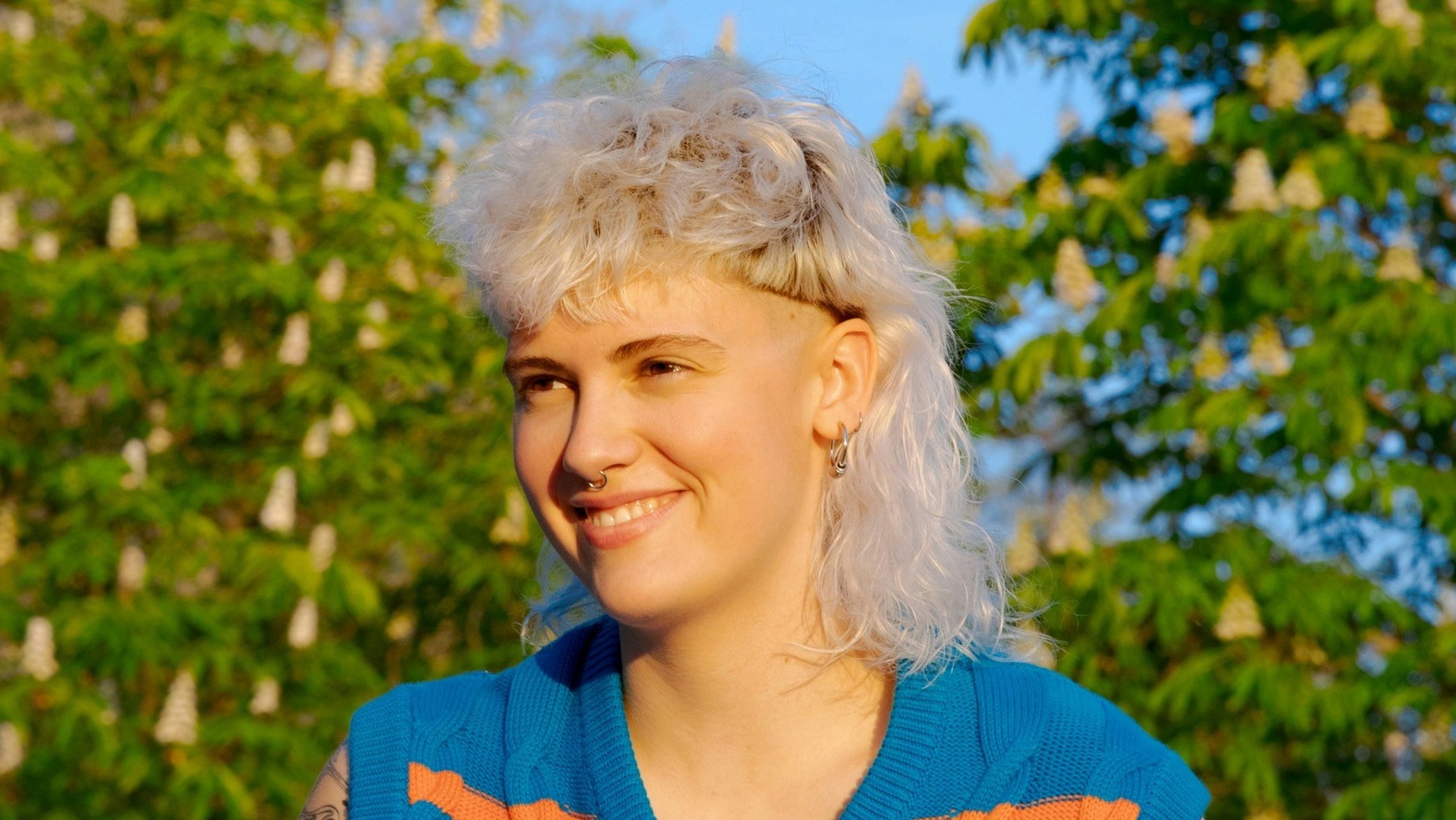 [ALT_TEXT: A young woman with a curly white blonde mullet haircut sitting outside on a sunny day.]
