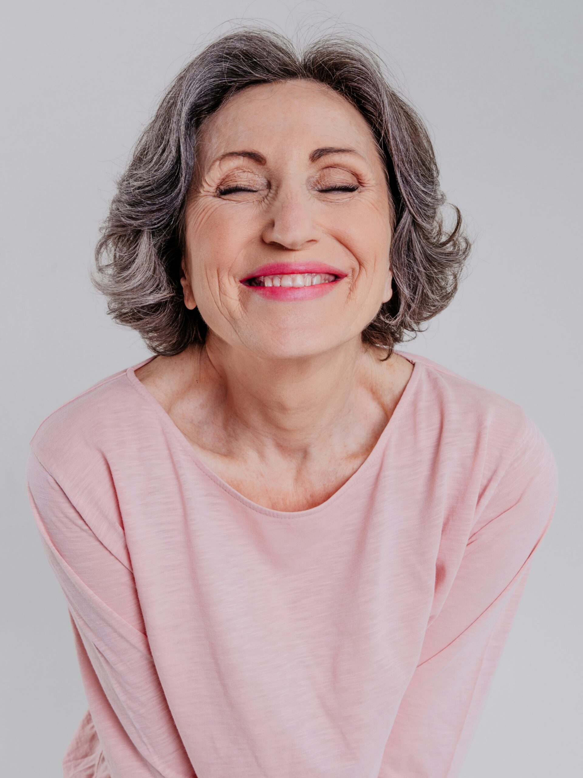 Portrait of a woman with short wavy salt-and-pepper hair wearing a loose pink top smiling with her eyes closed