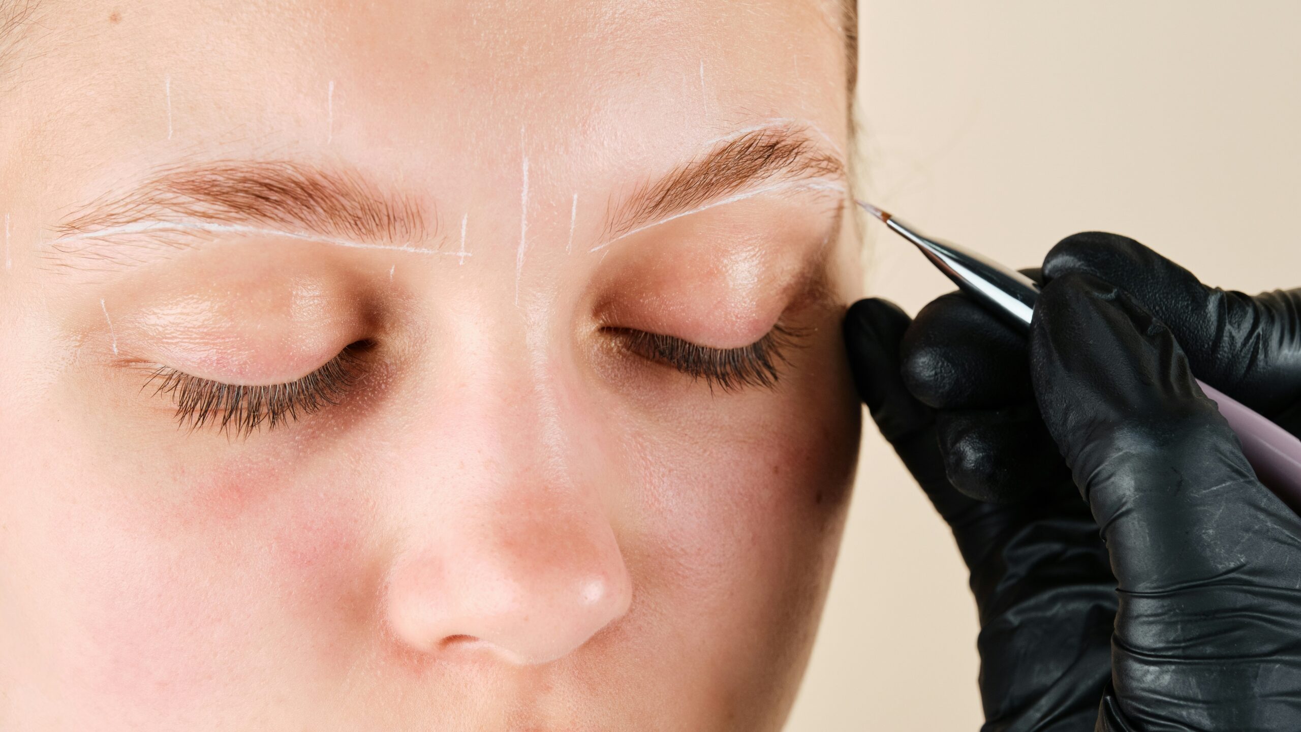 A close-up of a woman’s face as a technician draws white marks on her face to map the desired shape of her brows