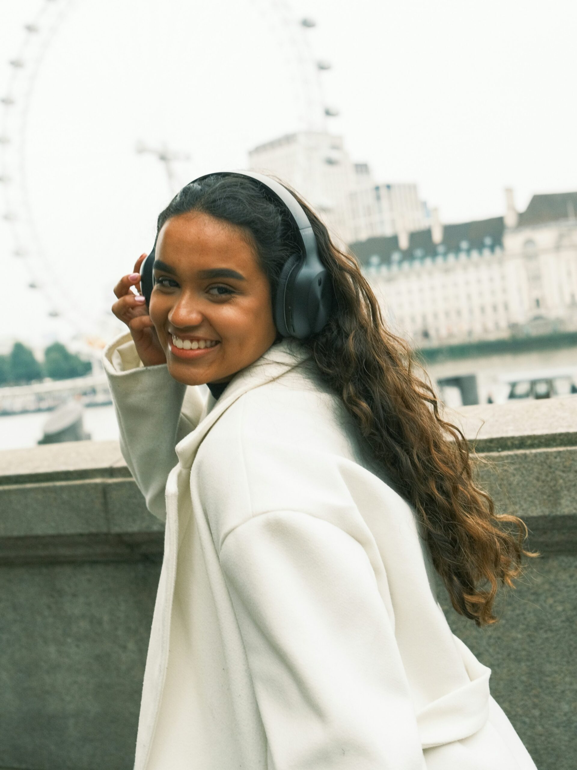 A smiling woman with microblading on her brows wearing black over-ear headphones and a white coat walking along the River Liffey in Dublin
