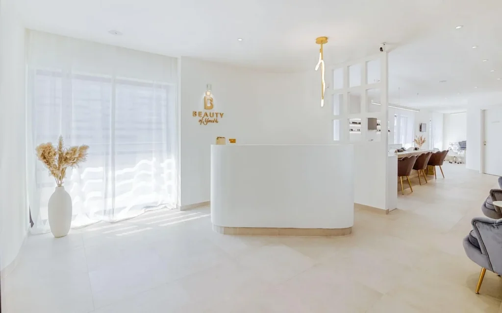A bright, all-white reception area with a curved desk, gold Beauty of Youth logo on the wall, a sculptural pendant light, sheer curtains filtering natural light, and brown velvet treatment chairs visible in the background.