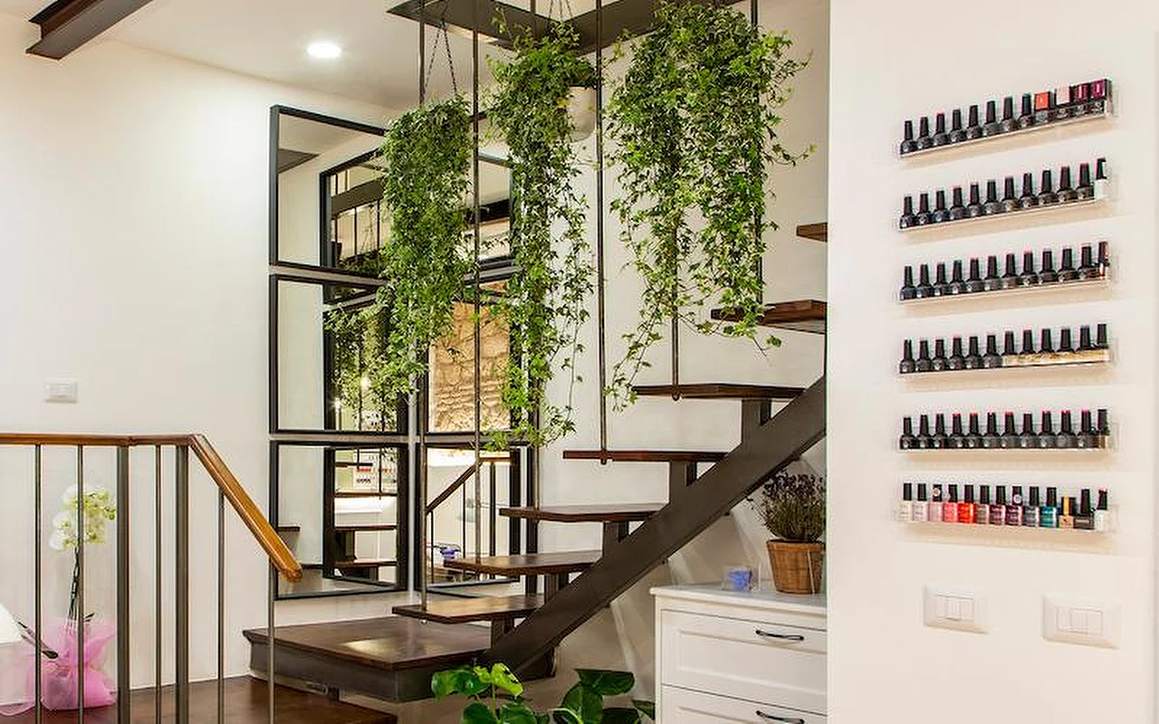 A stylish modern interior featuring a cantilevered wood and metal staircase. Lush trailing plants hang from the ceiling between the steps, and rows of colourful nail polish bottles are displayed on sleek transparent shelves on the white wall.