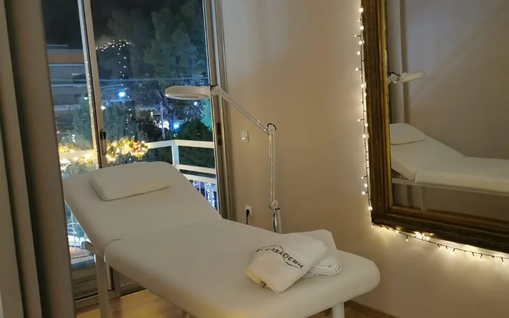 A softly lit treatment room with a white bed, a CasaDerm-branded towel, a magnifying lamp, fairy lights framing a gold mirror, and a balcony window looking out over the Glyfada evening streetscape.