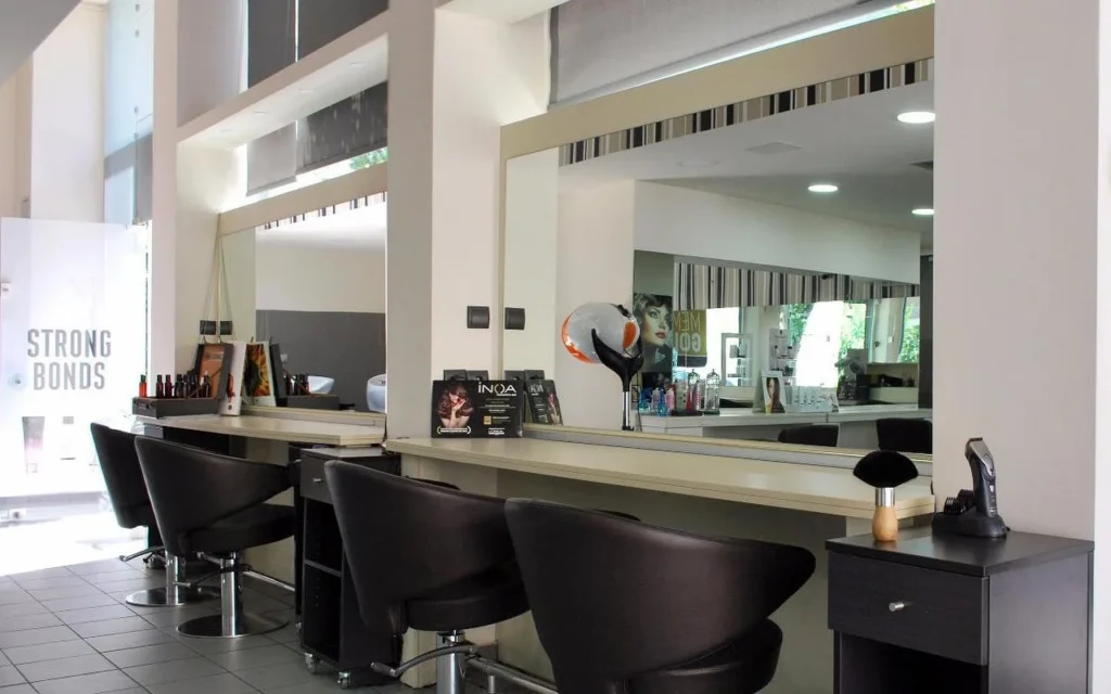 A professional hair salon with dark leather styling chairs facing large mirrors, hair products arranged along the counter, high ceilings with natural light filtering through transom windows.