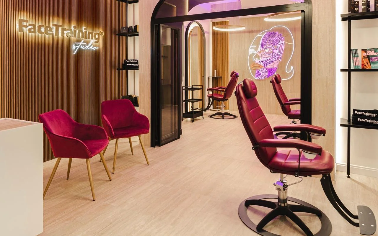 A minimalist studio featuring a wood-slatted feature wall with a backlit logo. The treatment area includes professional burgundy leather chairs and a neon light installation depicting a stylised facial muscle diagram on a glass partition.
