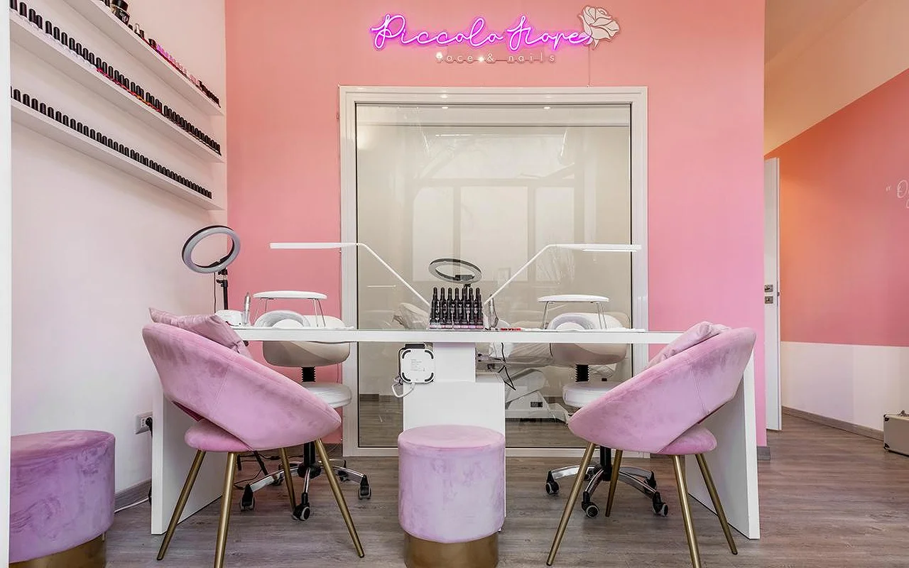 A vibrant manicure station with bright pink walls and a hot pink neon sign. Lilac velvet chairs with gold legs sit at a minimalist white desk equipped with professional LED nail lamps.