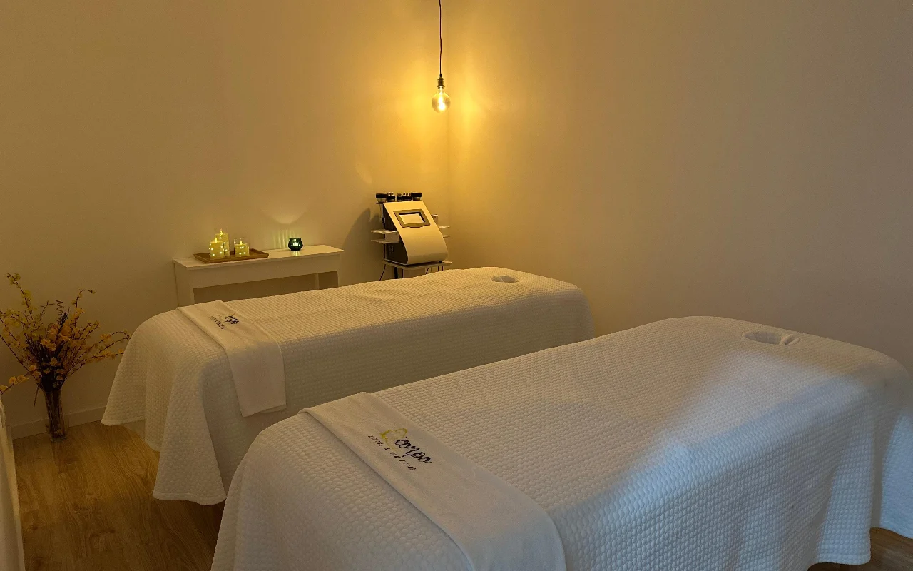 A tranquil massage or treatment room with two treatment tables placed side by side, dressed in crisp white towels and sheets. The room is illuminated by a single warm, exposed hanging light bulb and small candles on a side table, creating a serene atmosphere. A specialized cosmetic machine stands in the background between the beds.