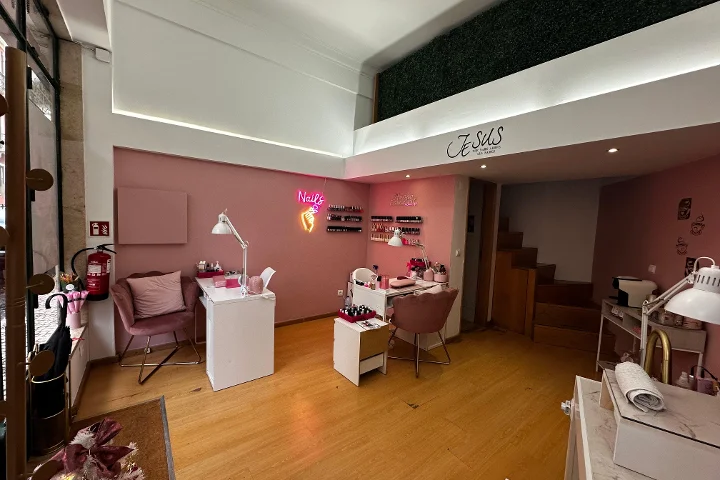 A chic nail studio with light pink walls and warm wooden floors. The room contains multiple white manicure desks paired with dusty pink velvet chairs. A bright pink neon sign shaped like a hand holding a nail polish bottle with the word 'Nails' glows on the wall, and rows of nail polishes are displayed on small shelves. A mezzanine floor with a green faux-hedge wall is visible above.