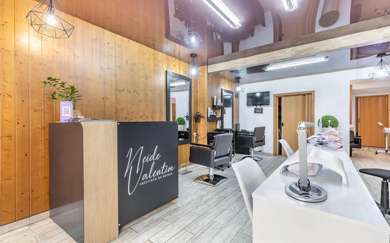 A contemporary beauty salon featuring a reception desk on the left branded with 'Neide Valentim Instituto de Beleza'. The walls are finished with warm vertical wooden panelling and large mirrors. Black styling chairs stand in front of the mirrors, while a white manicure station with white chairs runs along the right side of the grey tiled floor.