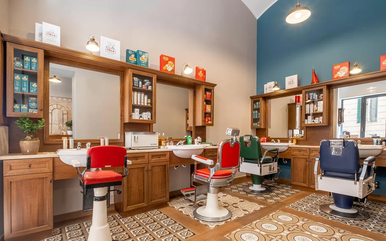 A high-end traditional barbershop with rich wooden cabinetry, vintage-style patterned floor tiles, and professional barber chairs in red, green, and navy blue. Traditional grooming products are displayed on the shelving units.