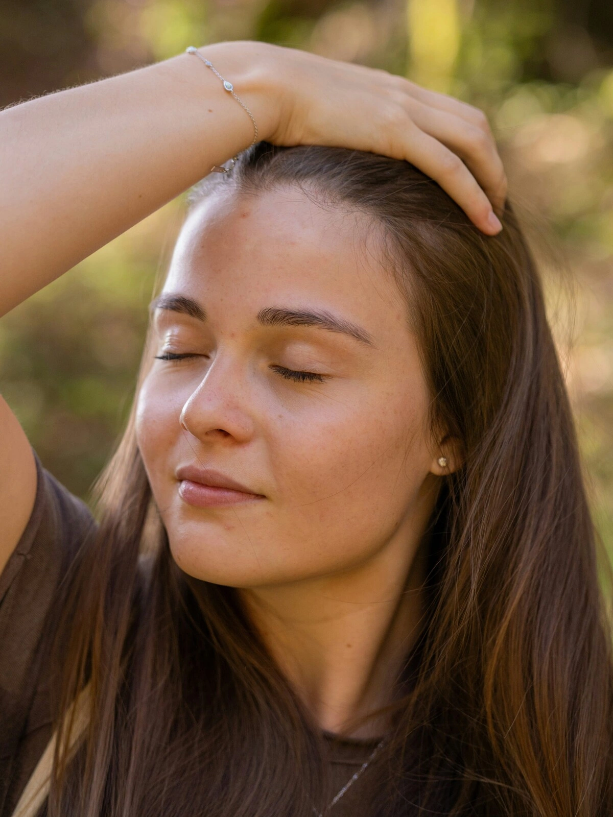 A woman with her eyes closed and a relaxed expression running her hand over her head and through her long brown a hair