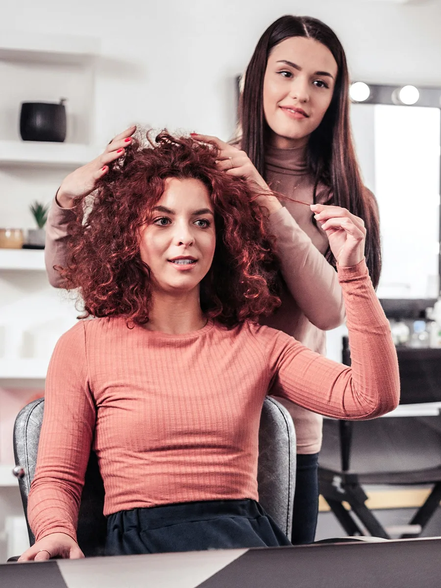 A client with dyed red curly hair sits at the hairdressers while her stylist discusses her treatment