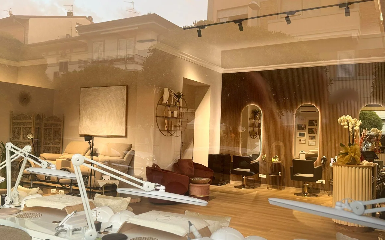 The interior of Lilian Almanza’s salon seen through the shop window at golden hour. Nail stations with adjustable lamps line the left side, while the right features warm wood panelling, arched mirrors with backlit frames, and velvet armchairs.