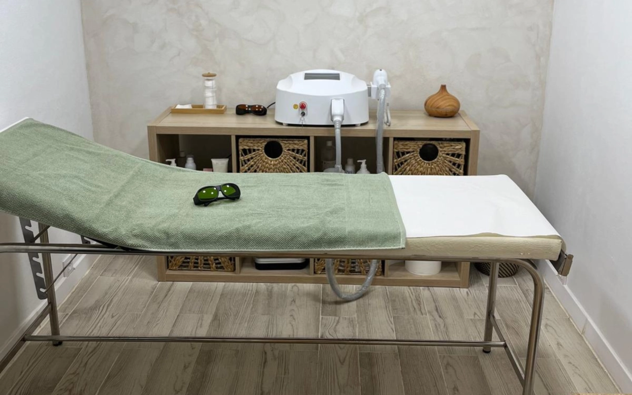 A neutral-toned treatment room with a metal-framed bed draped in a sage-green towel, a pair of dark protective goggles resting on top, a laser machine on a wooden console, and woven storage baskets below.