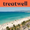 An aerial view of Playa de Palma in Mallorca, with a long curve of golden sand, rows of beach umbrellas, calm turquoise water, and a palm-lined promenade running alongside the city, with hills in the distance under a clear blue sky.