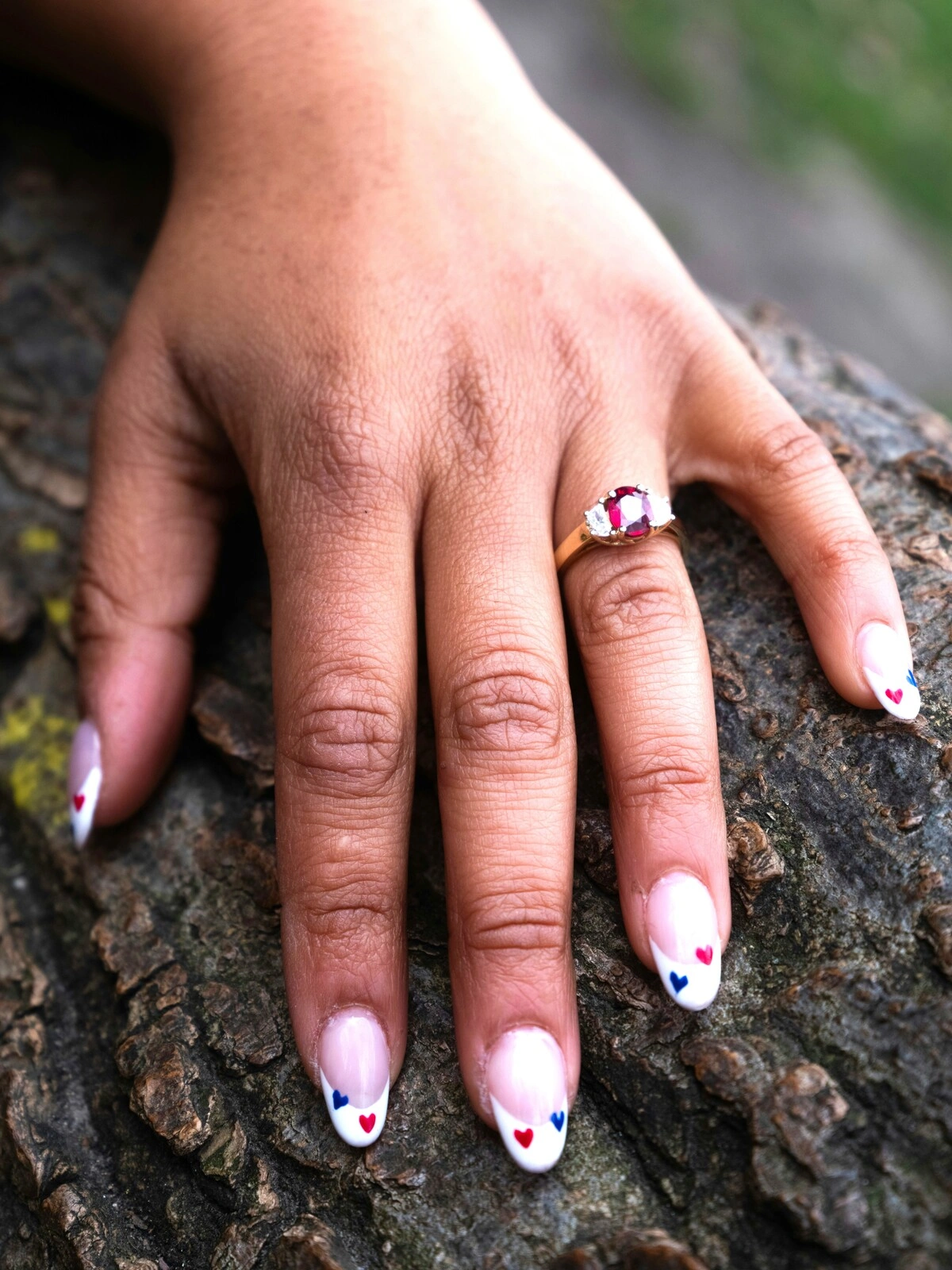A woman’s hand rests on a tree branch showing a French tips manicure on oval nails embellished with small red and blue hearts