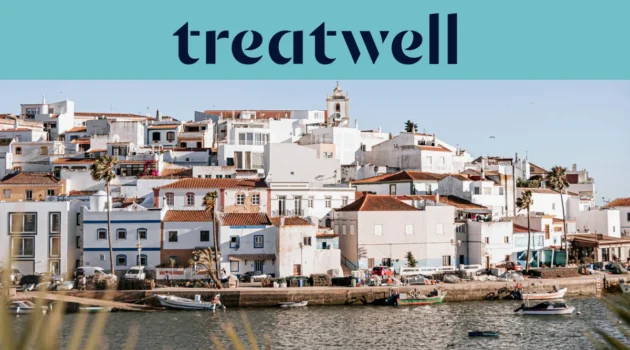 A whitewashed Algarve fishing village of terracotta-roofed houses rising up a hillside above a calm waterway, with small fishing boats moored along the quay, palm trees and a church tower under a clear blue sky.