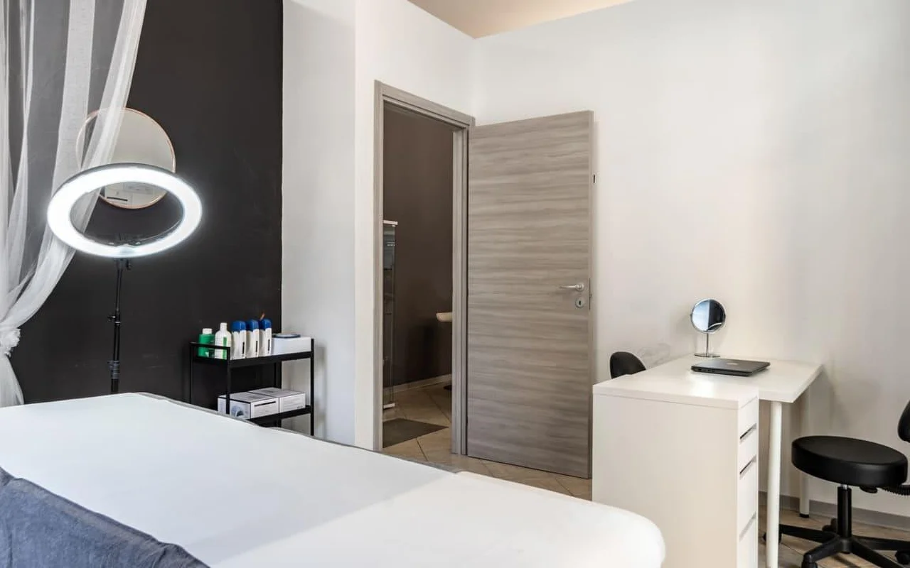 A treatment room at Coquette Wellness and Beauty, with a white treatment bed, an illuminated ring light on a stand against a black feature wall, sheer curtains at the window and a small white desk with a vanity mirror beside a doorway to an ensuite.