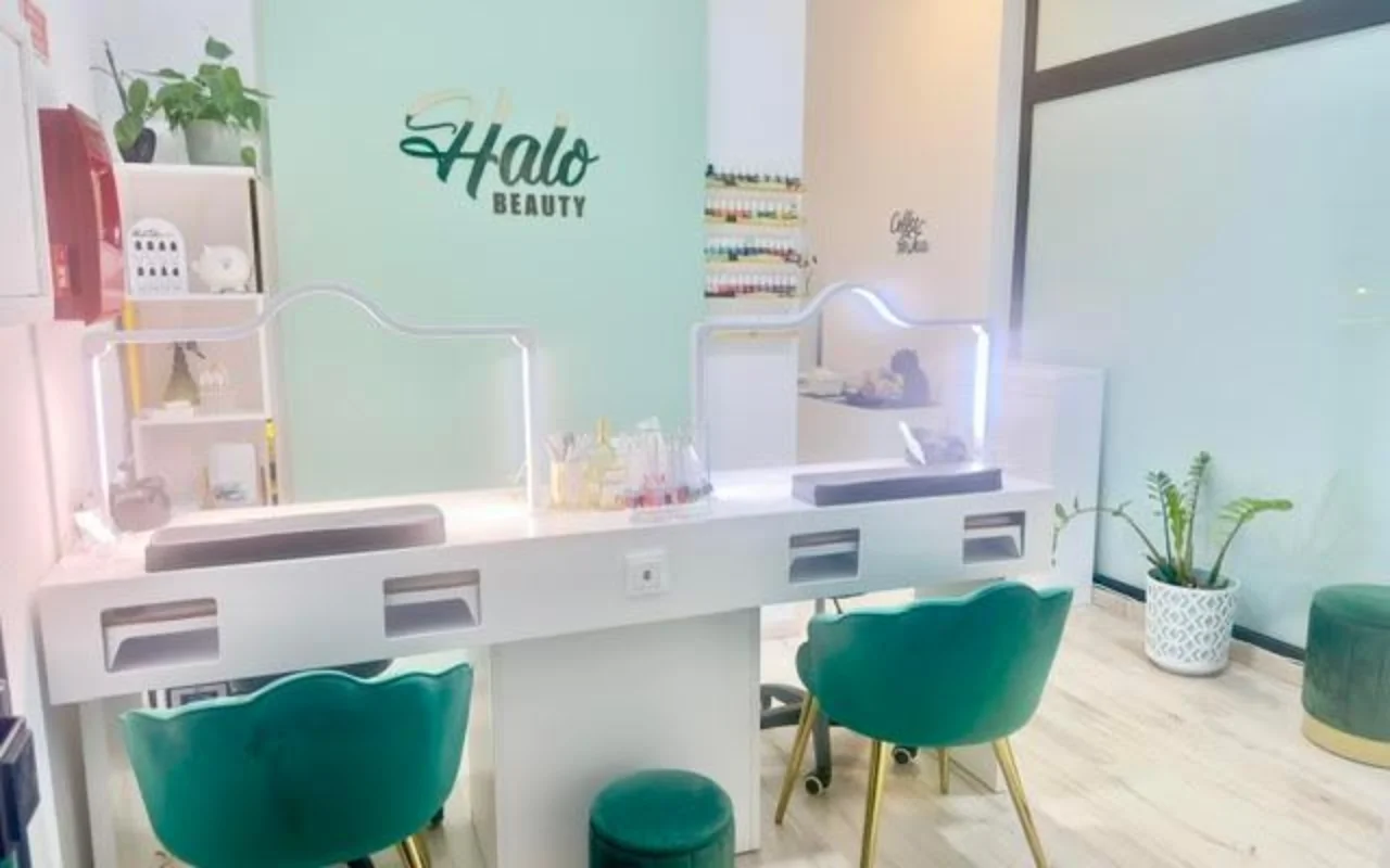 A nail salon with a mint-green feature wall carrying the Halo Beauty logo, a white double manicure desk with lit perspex screens, two emerald-green velvet tub chairs, a wall rack of nail polishes and potted plants.