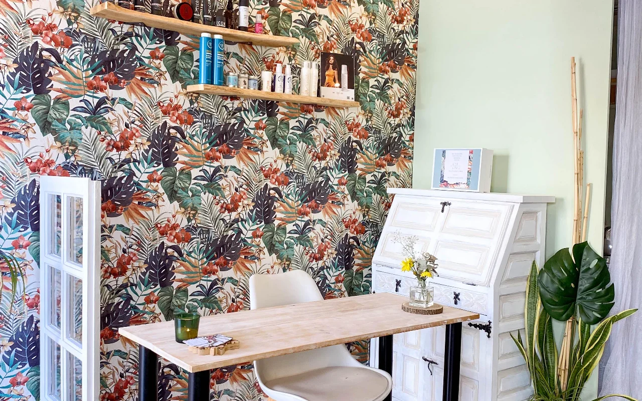 A styling station at Shangara Lora Organics in Marbella, with bold tropical wallpaper, a wooden table, a white antique bureau and organic haircare on floating shelves.
