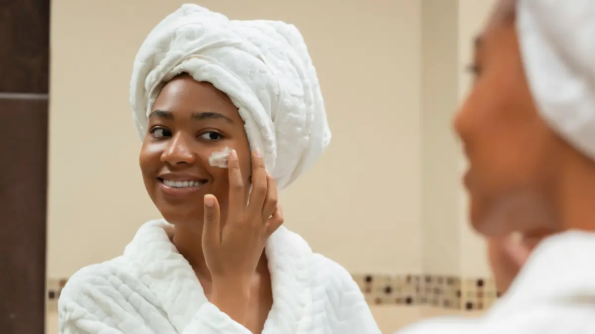 A woman wearing a white robe and a white towel wrapped around her head smiles in the bathroom mirror as she applies SPF to her face