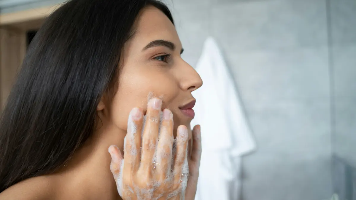 Side view of a woman massaging a cleanser into her face as she bends over a bathroom sink