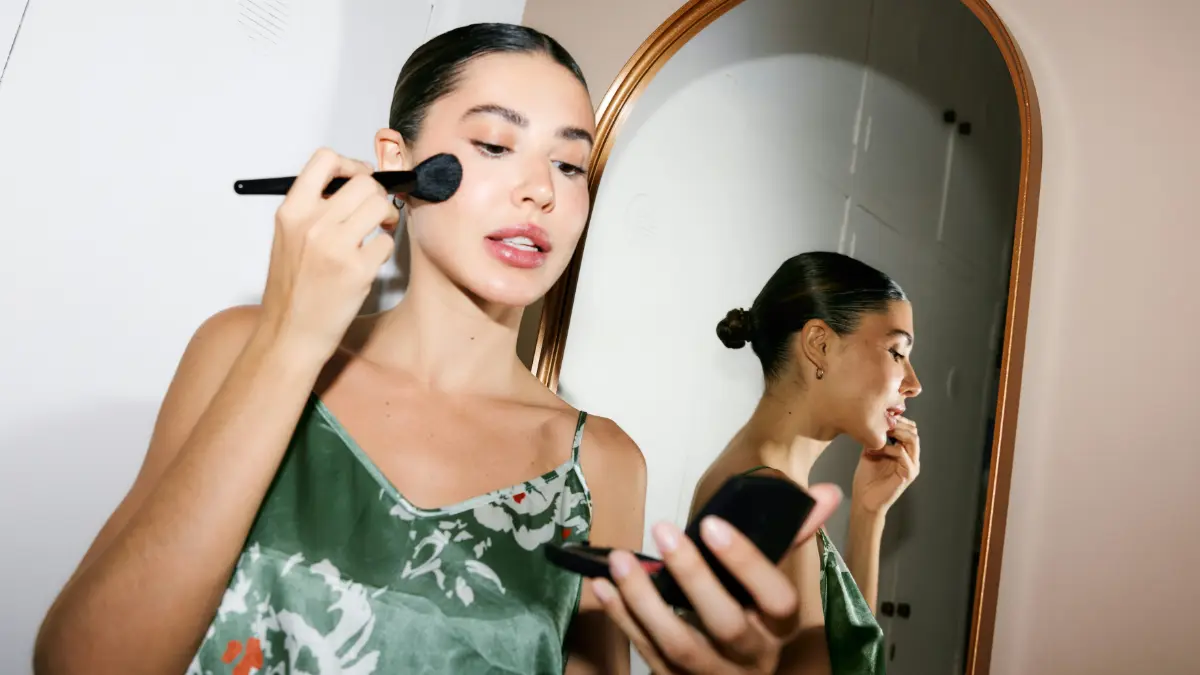 A glamorous woman dressed ready to go out looks into a pocket mirror as she applies makeup with a brush
