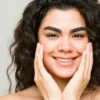 A young woman with clear skin and dark wavy hair smiles at the camera