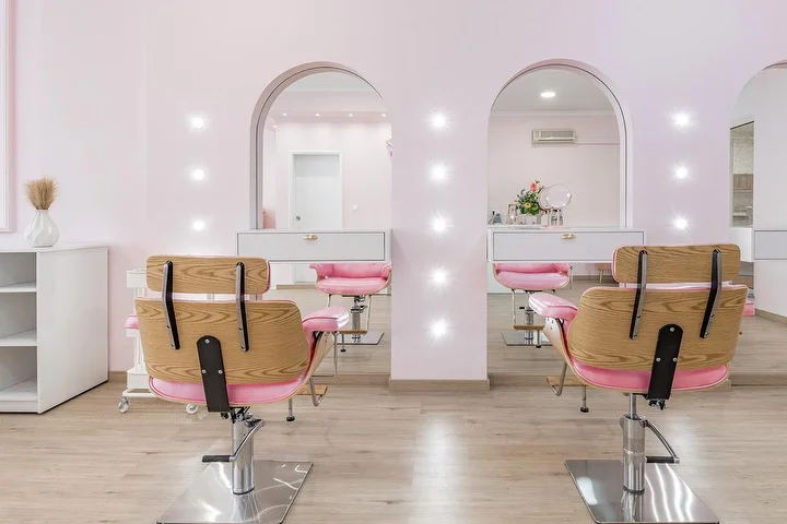 A bright hair salon in soft pink, with wooden and pink upholstered styling chairs facing two arched, spot-lit mirror alcoves, white cabinets and pale wood-effect flooring.