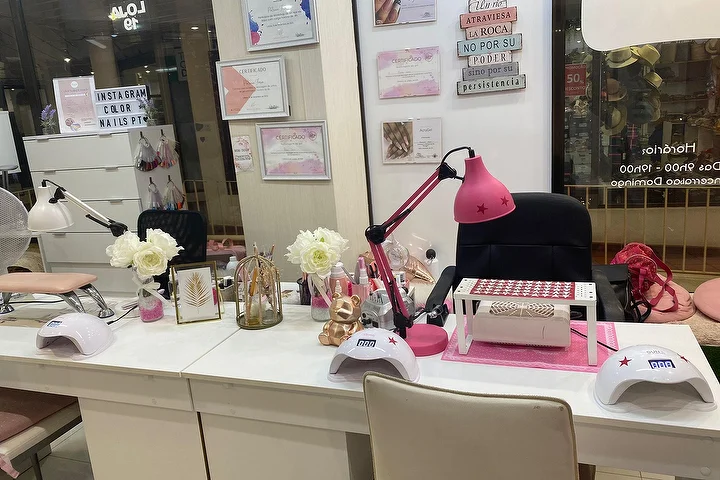 A nail salon station with white desks, a bright pink lamp and LED nail dryers, white roses and framed certificates on the wall, and the Color Nails PT name shown on the glass alongside the opening hours.