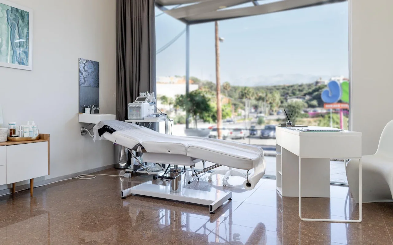A clean white treatment room with a reclining beauty bed, skincare equipment on a trolley, framed artwork and a large window looking out over trees, a road and distant hills.