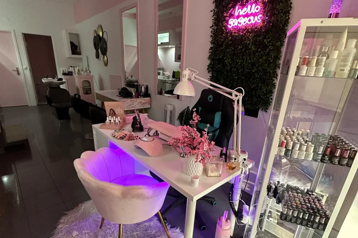 A pink-lit beauty station with a neon 'hello gorgeous' sign on a foliage wall, a cream shell-shaped chair, a white desk with brushes and pink blossom, and a glass cabinet of colourful nail polishes.