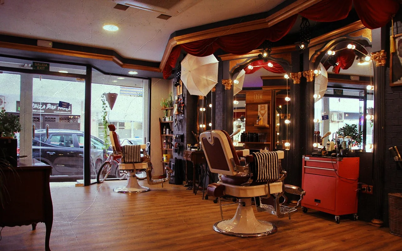 A vintage-style barbershop with classic leather reclining chairs on chrome bases and striped towels. Gilded mirrors framed by round bulb lights and red velvet drapery line the wall, a red tool trolley stands to one side, and street-facing windows light the wooden floor.