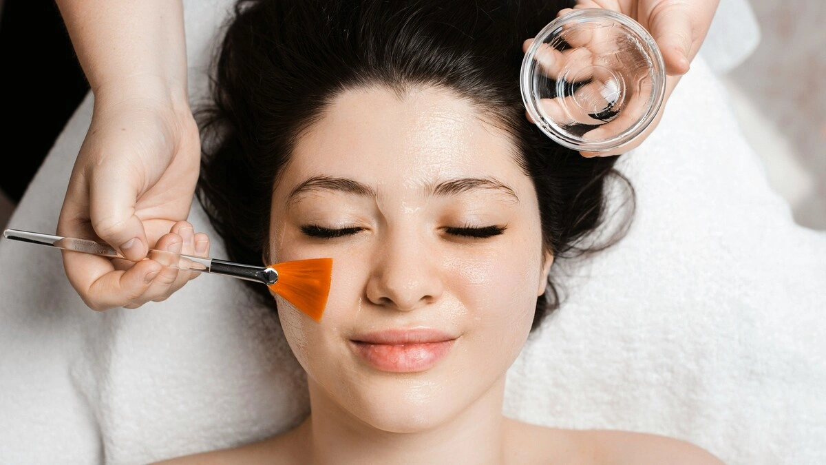Top view of an aesthetician applying a chemical peel solution to a client’s face using a wide, flat brush