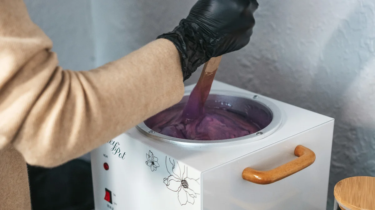 A therapist’s hand wearing a black glove stirs a pot of melted hot wax at a beauty salon