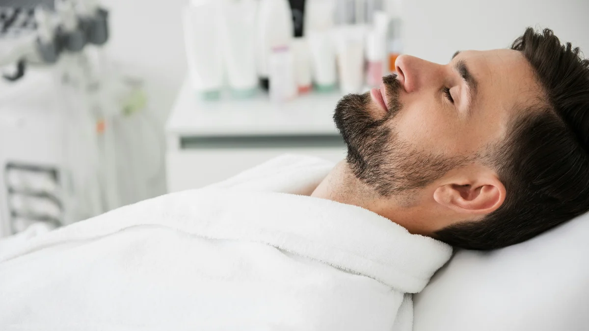 A man lays on a treatment bed wearing a white robe and rests his head back with his eyes closed