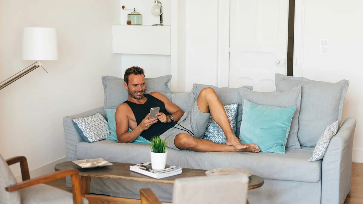 A man relaxes with his feet up on his sofa smiling as he checks his phone, he wears a black vest top and loose, soft grey shorts