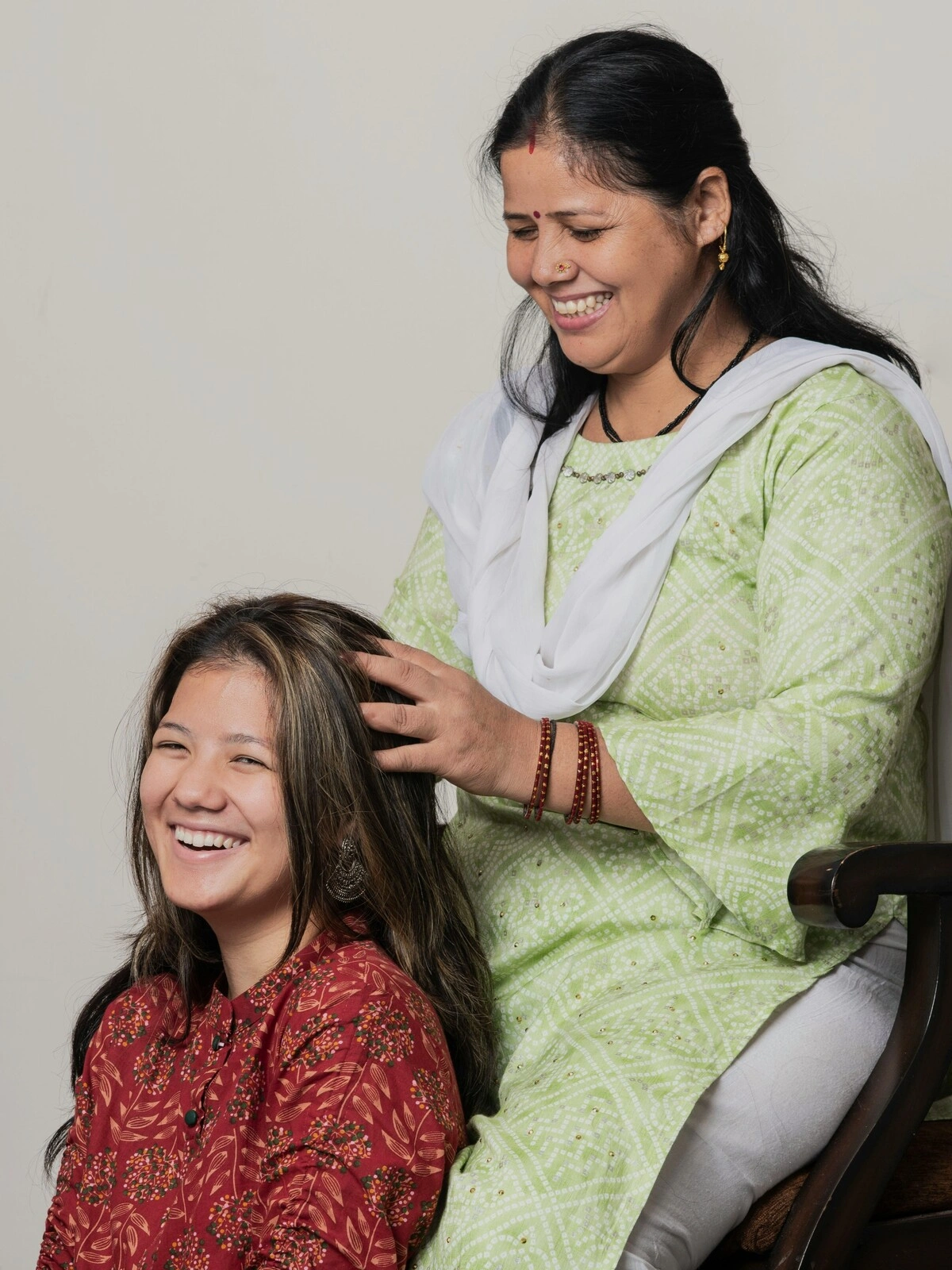 An Indian woman sitting in a wooden chair wearing traditional dress performs an Indian head massage on a woman seated on the floor